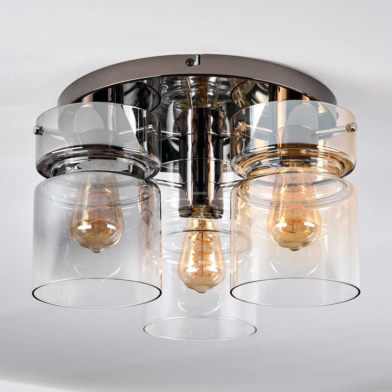 Eine moderne Deckenleuchte mit drei Glasschirmen, jede mit einer warm getönten Glühbirne. Das Gerät hat ein metallisches Finish und ist an einer weißen Decke montiert.