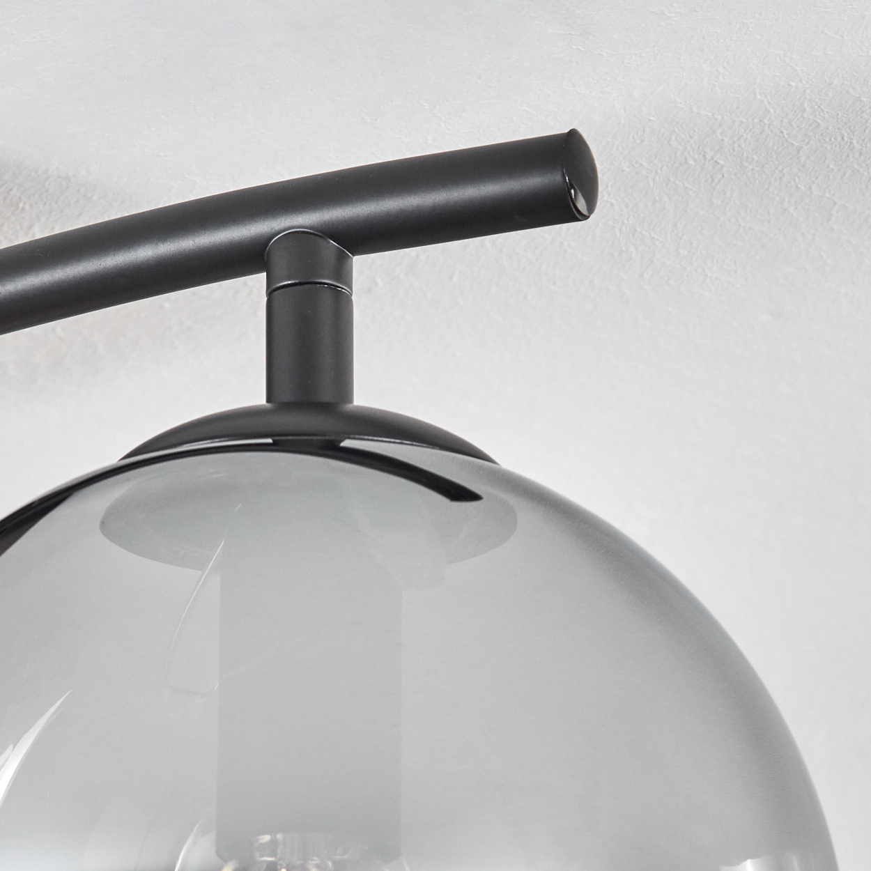 Nahaufnahme einer schwarzen Lampenhalterung mit rauchfarbenem Glasschirm. Die Halterung befindet sich vor einer strukturierten weißen Wand.