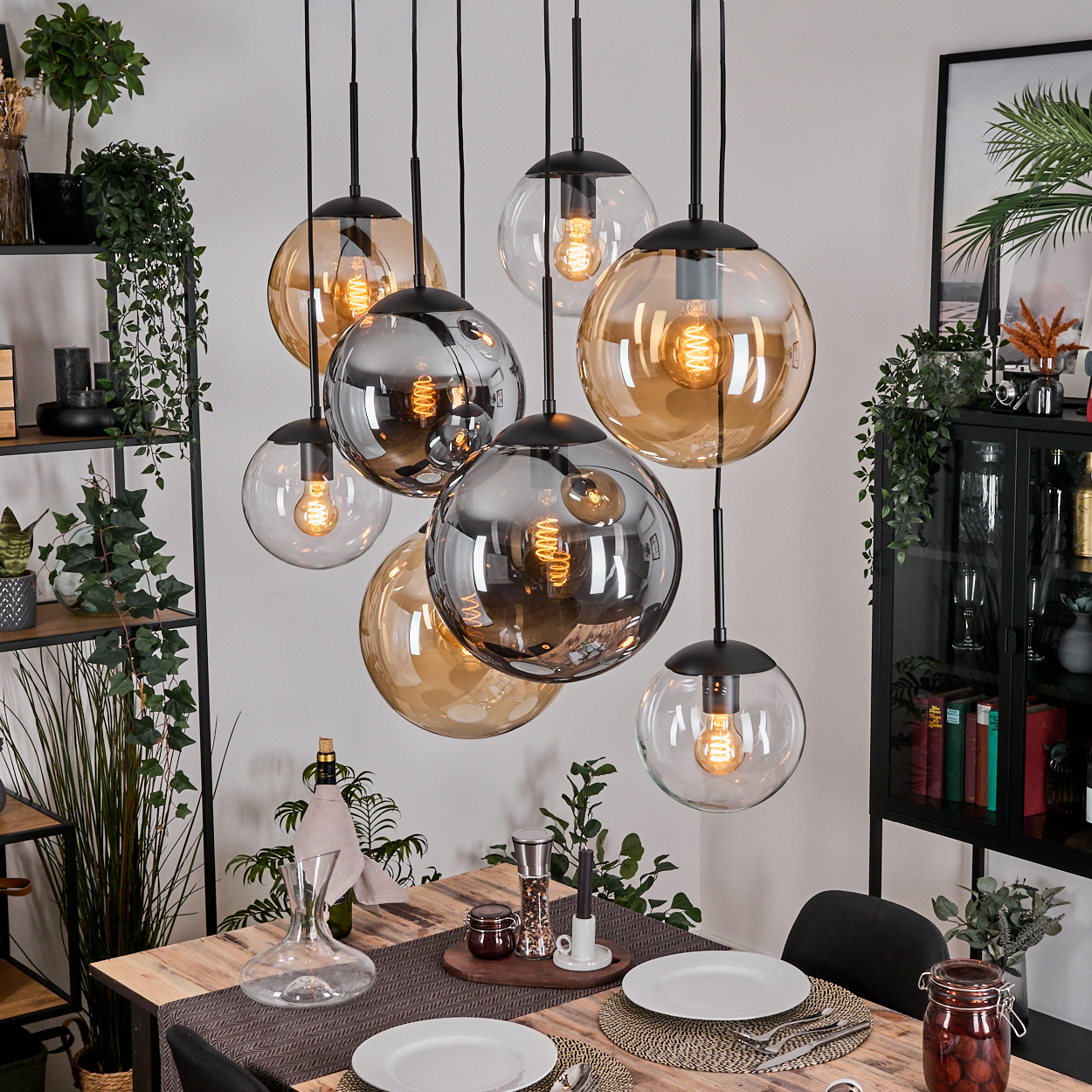 Esszimmer mit Holztisch, Glaskugellampen und Pflanzen. Teller und Besteck sind gedeckt. Karaffe und Gläser sind vorhanden.