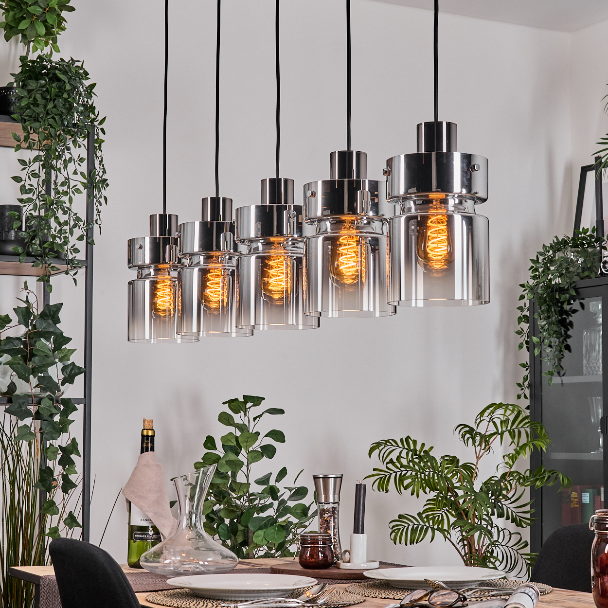 Esszimmer mit Hängelampen, Pflanzen und gedecktem Tisch. Lampen haben freiliegende Glühbirnen. Weitere Elemente: Wein, Dekanter und Geschirr.