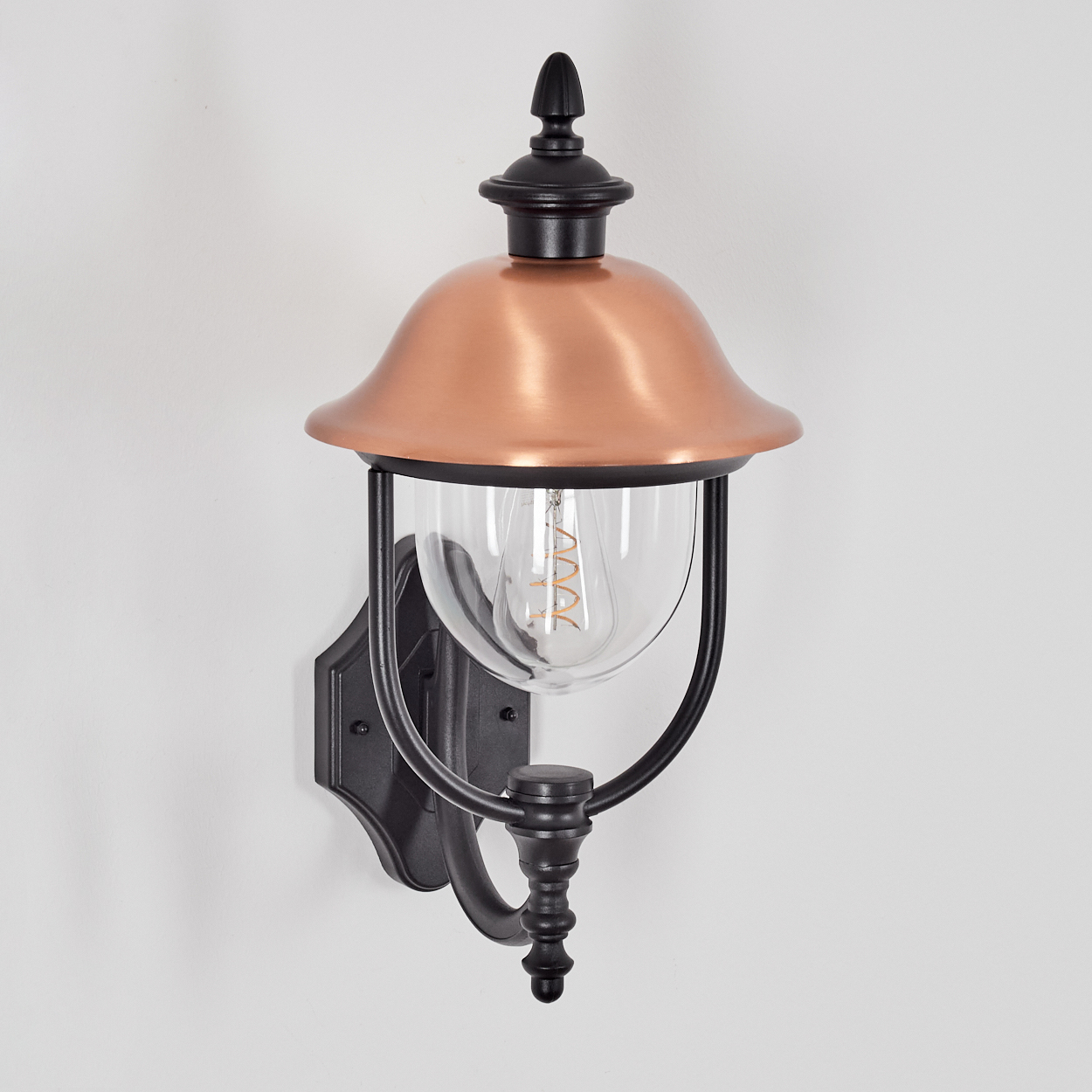 Eine an der Wand montierte Außenlampe mit kupferfarbenem Schirm, klarem Glas und schwarzem Rahmen. Sie ist an einer weißen Wand befestigt.