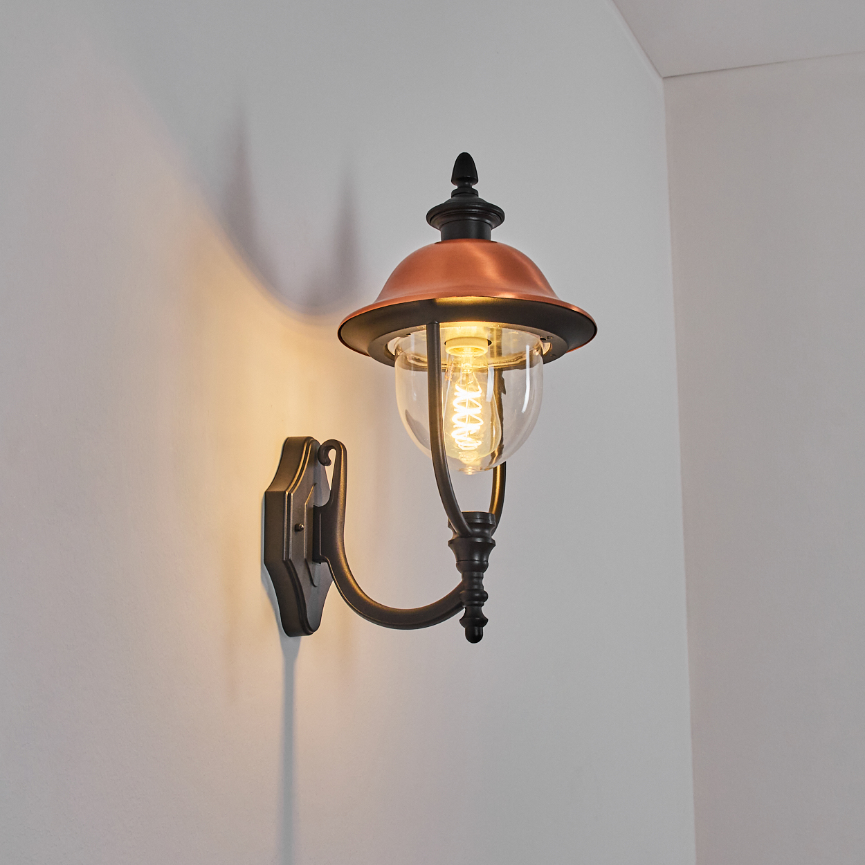 Eine Wandlaterne mit kupferfarbener Oberseite und Glaskugel, die ein warmes Licht und Schatten wirft.