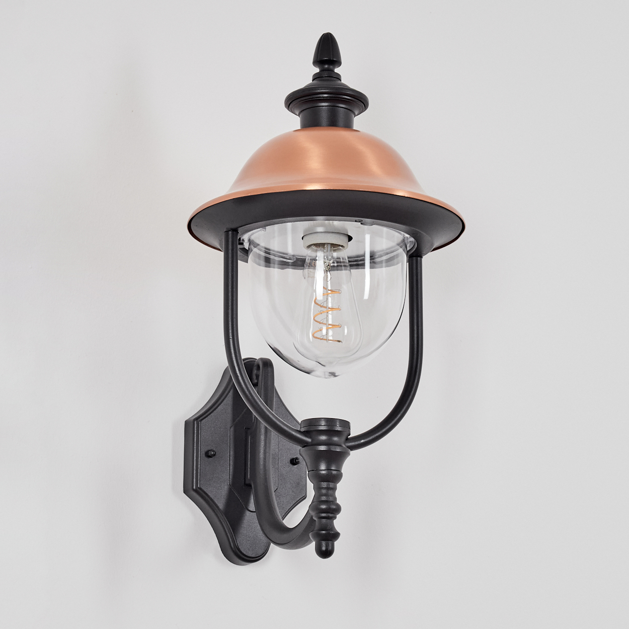 Eine dekorative Wandlampe mit kupferfarbenem Oberteil und einem klaren Glaskörper für die Glühbirne. Die Lampe ist schwarz.