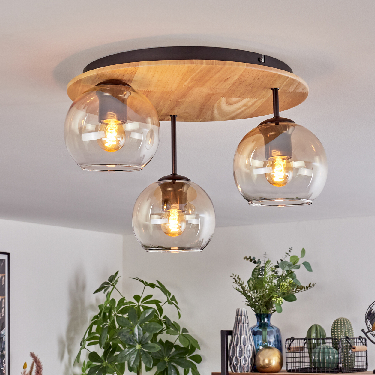 Moderne Deckenlampe mit drei Hängelampen. Jede hat einen kugelförmigen, bernsteinfarbenen Glasschirm. Die Umgebung umfasst Pflanzen und Vasen.