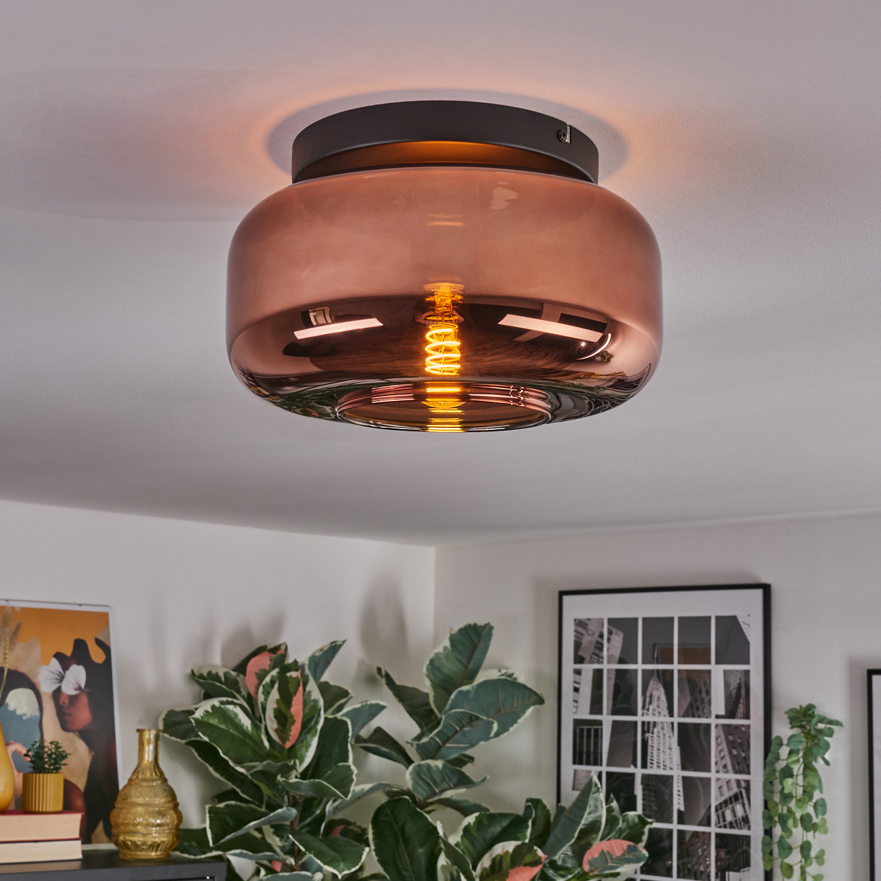 Kupferfarbene Deckenlampe mit sichtbarer Glühbirne. Pflanzen und gerahmte Kunstwerke sind im Hintergrund sichtbar.