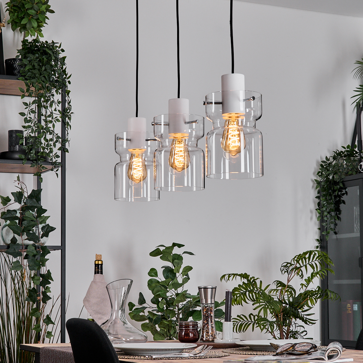 Drei Pendelleuchten mit Edison-Glühbirnen hängen über einem Tisch, der mit Pflanzen und einer Karaffe gedeckt ist.