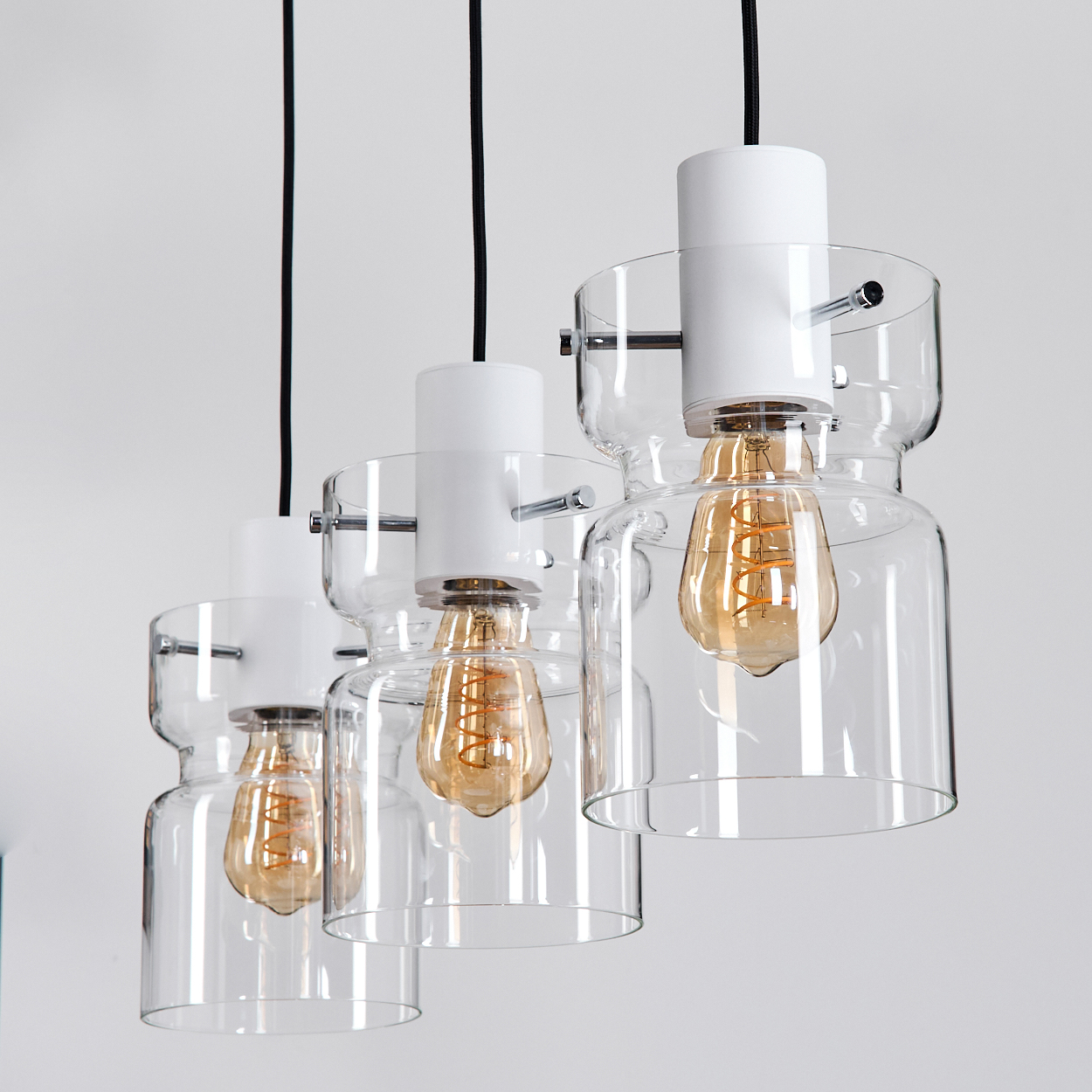 Drei Pendelleuchten mit Glasschirmen und Edison-Glühbirnen hängen vor einer weißen Wand. Die Leuchten haben einen weißen Aufsatz und einen Glasboden.