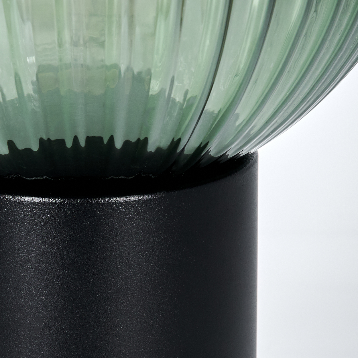 Nahaufnahme einer Lampe. Ein geriffelter, grüner Glasschirm steht auf einem schwarzen, zylindrischen Sockel. Weißer Hintergrund.