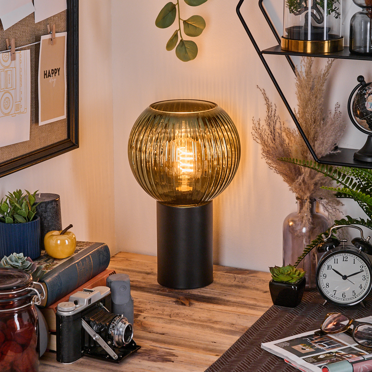 Ein Schreibtisch mit Lampe, Büchern, Kamera und Uhr. Eine Pinnwand hängt an der Wand. Regale halten Pflanzen und Deko.
