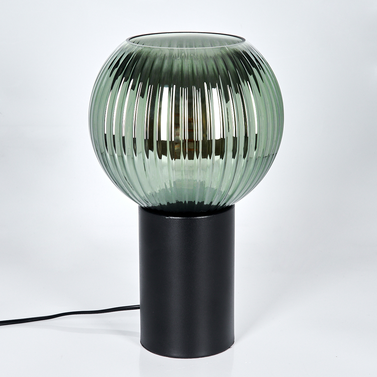 Eine moderne Lampe mit grünem, geripptem Glasschirm steht auf einem schwarzen, zylindrischen Sockel. Ein schwarzes Netzkabel geht ab.