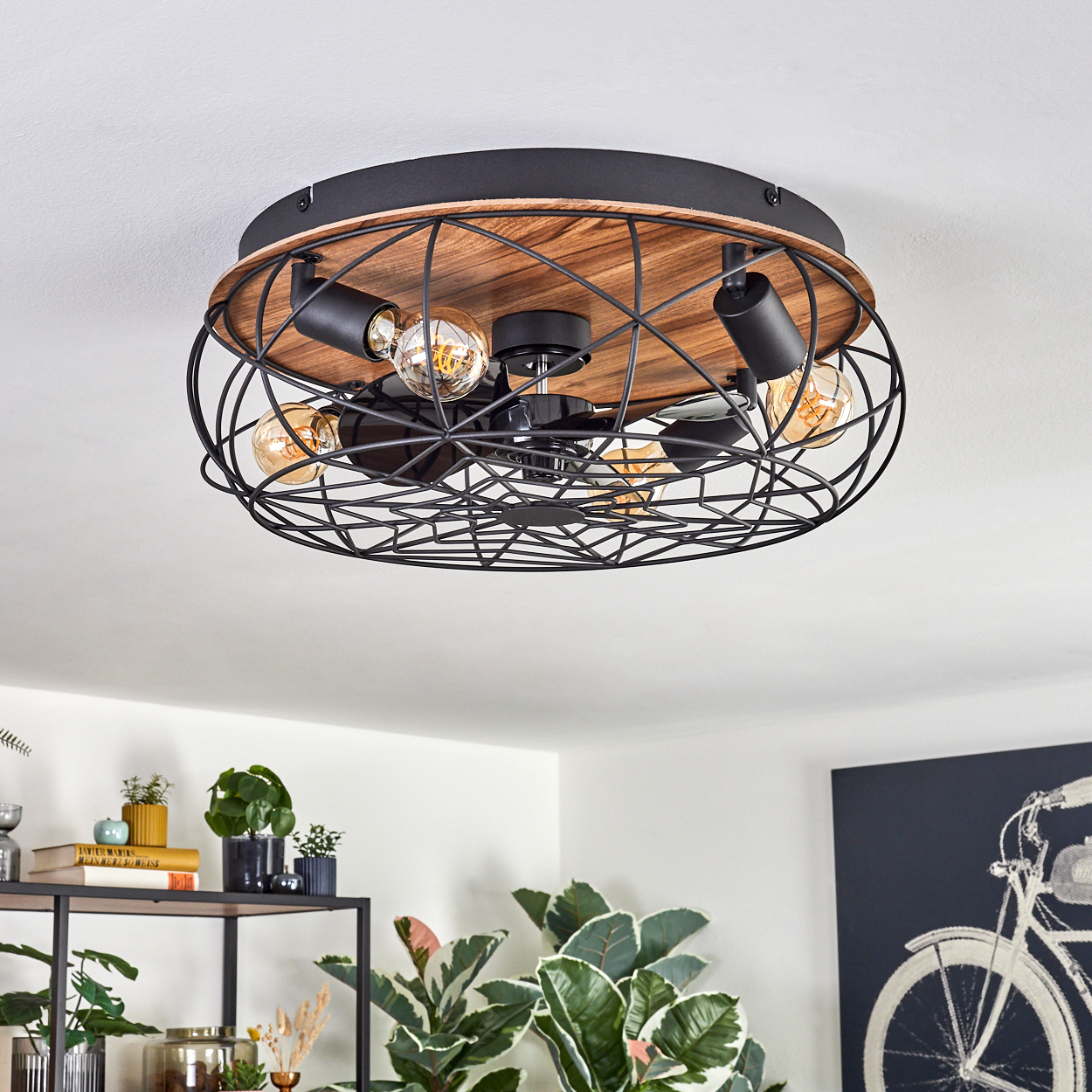 Deckenleuchte mit Holzsockel, schwarzem Metallkäfig und freiliegenden Glühbirnen. Pflanzen und ein Fahrradkunstwerk sind unten sichtbar.