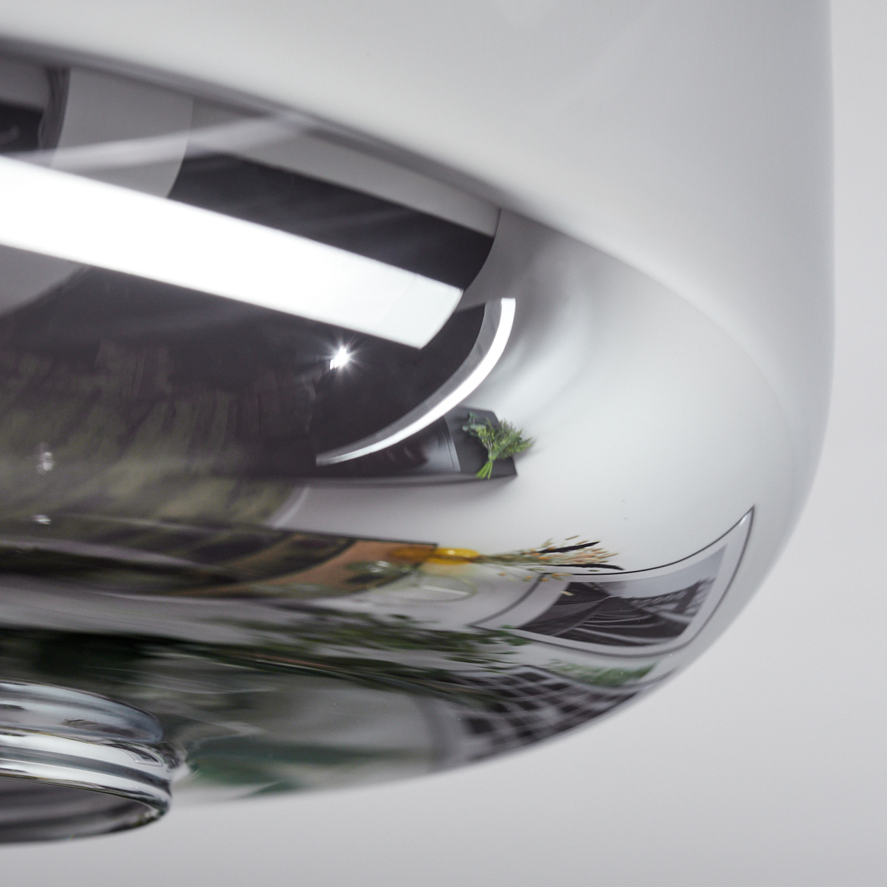 Nahaufnahme einer reflektierenden, chromartigen Oberfläche. Sie spiegelt einen dunklen Innenraum mit einem Regal mit grünen Pflanzen wider.