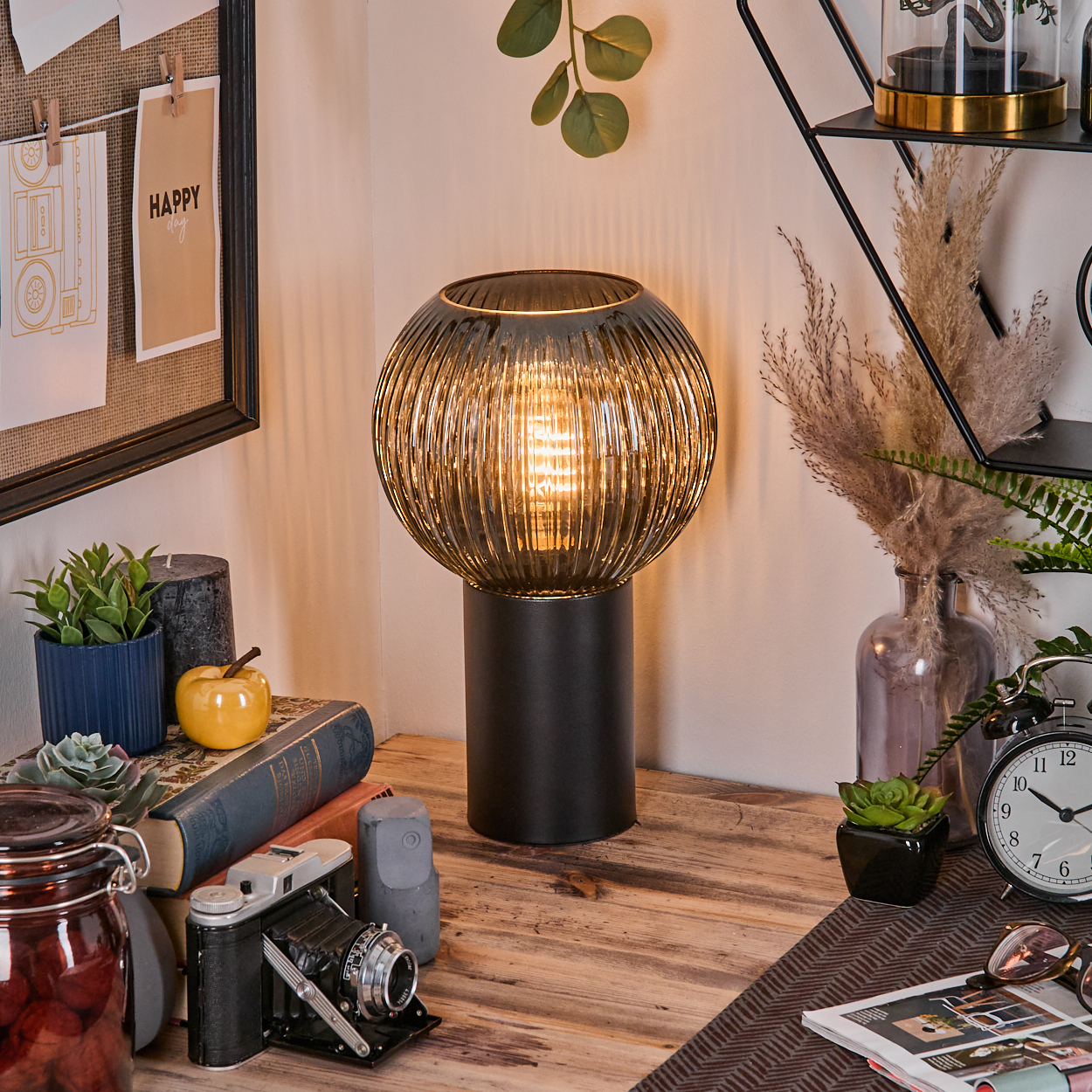 Ein Schreibtisch mit Lampe, Büchern, Kamera und Pflanzen. Eine Pinnwand mit 'Happy'-Notiz. Ein Glas mit roten Gegenständen links.