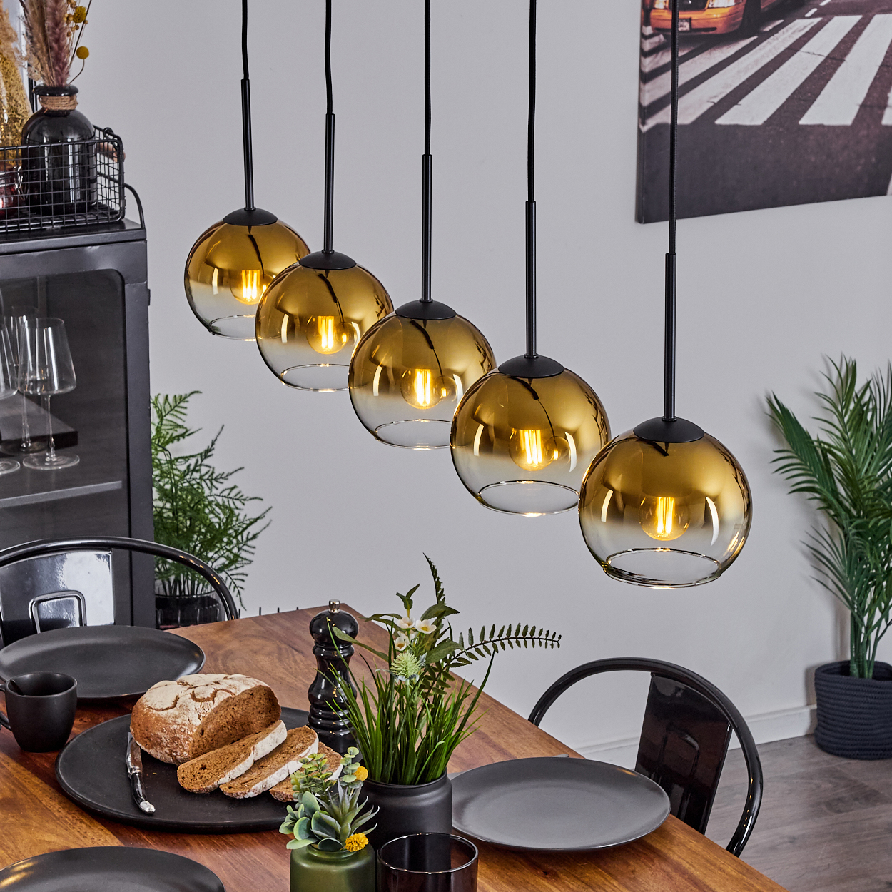 Esstisch mit Brot, Tellern und hängenden goldenen Lichtern. Im Hintergrund ein Schrank, Gemälde und Pflanzen.