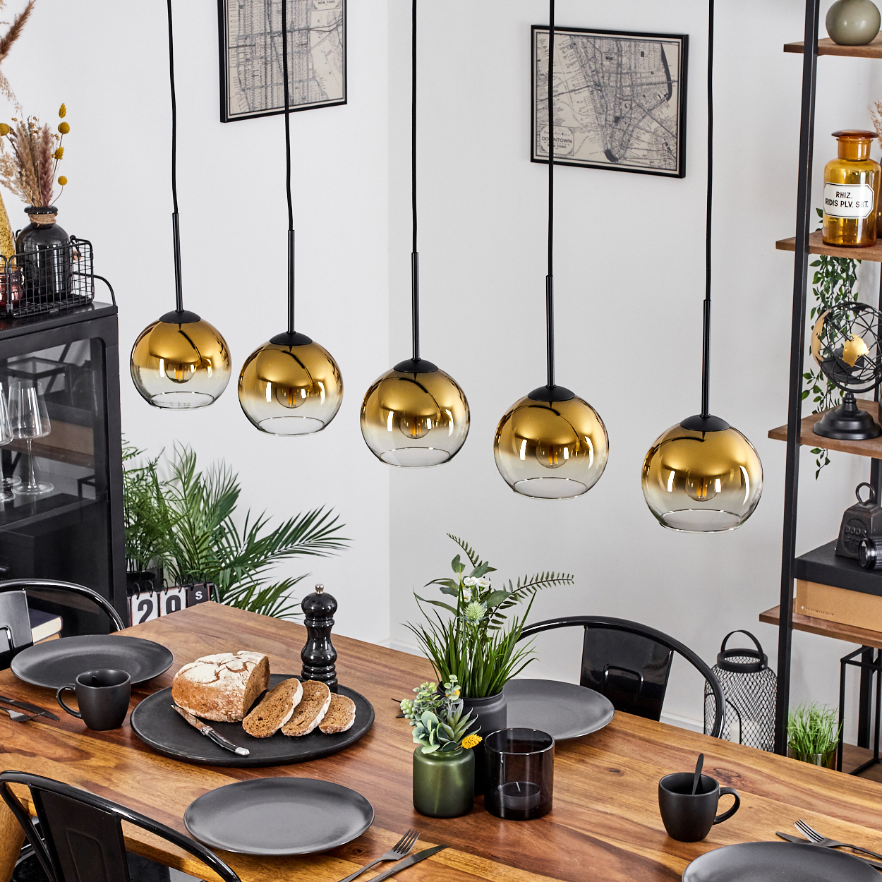 Esszimmer mit Holztisch, fünf goldenen Pendelleuchten, Brot, Tellern und Pflanzen. Schwarze Stühle sind sichtbar.