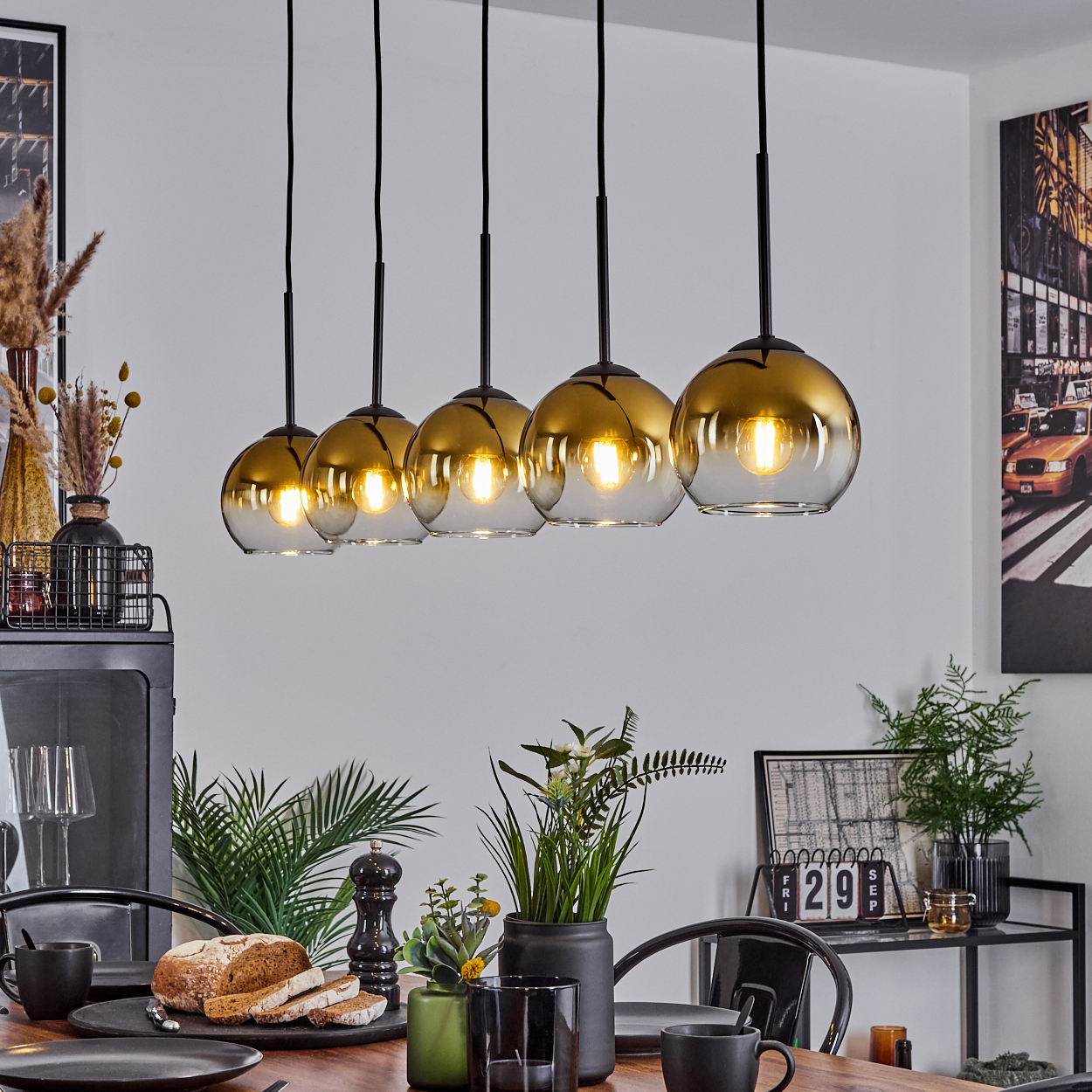 Esszimmer mit hängenden gold getönten Kugelleuchten. Tisch mit Brot, Pflanzen und schwarzem Geschirr. Kunst an den Wänden.