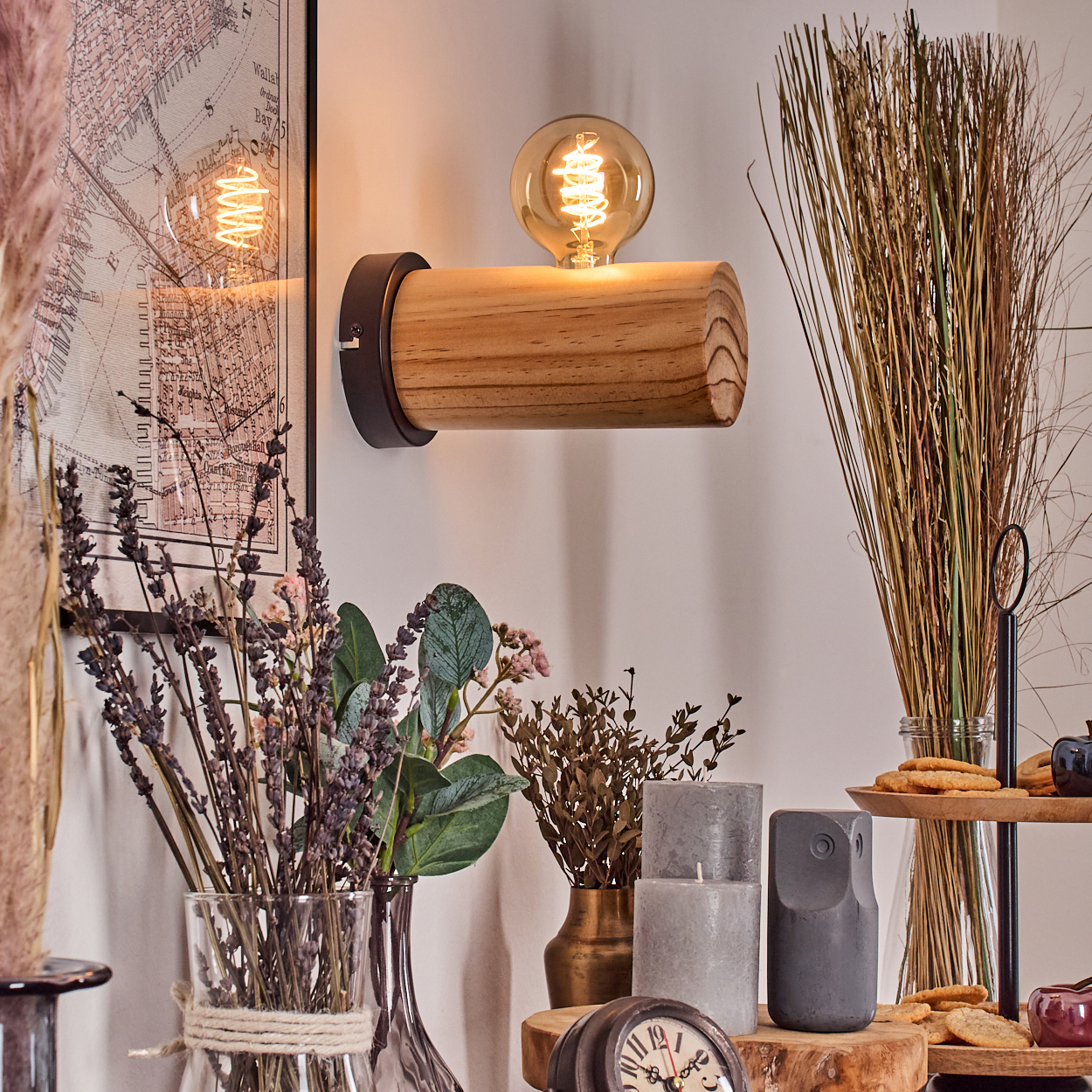 Eine hölzerne Lampe mit einer leuchtenden Glühbirne. Trockenblumen und Kerzen auf einem Tisch. Eine Karte hängt an der Wand.