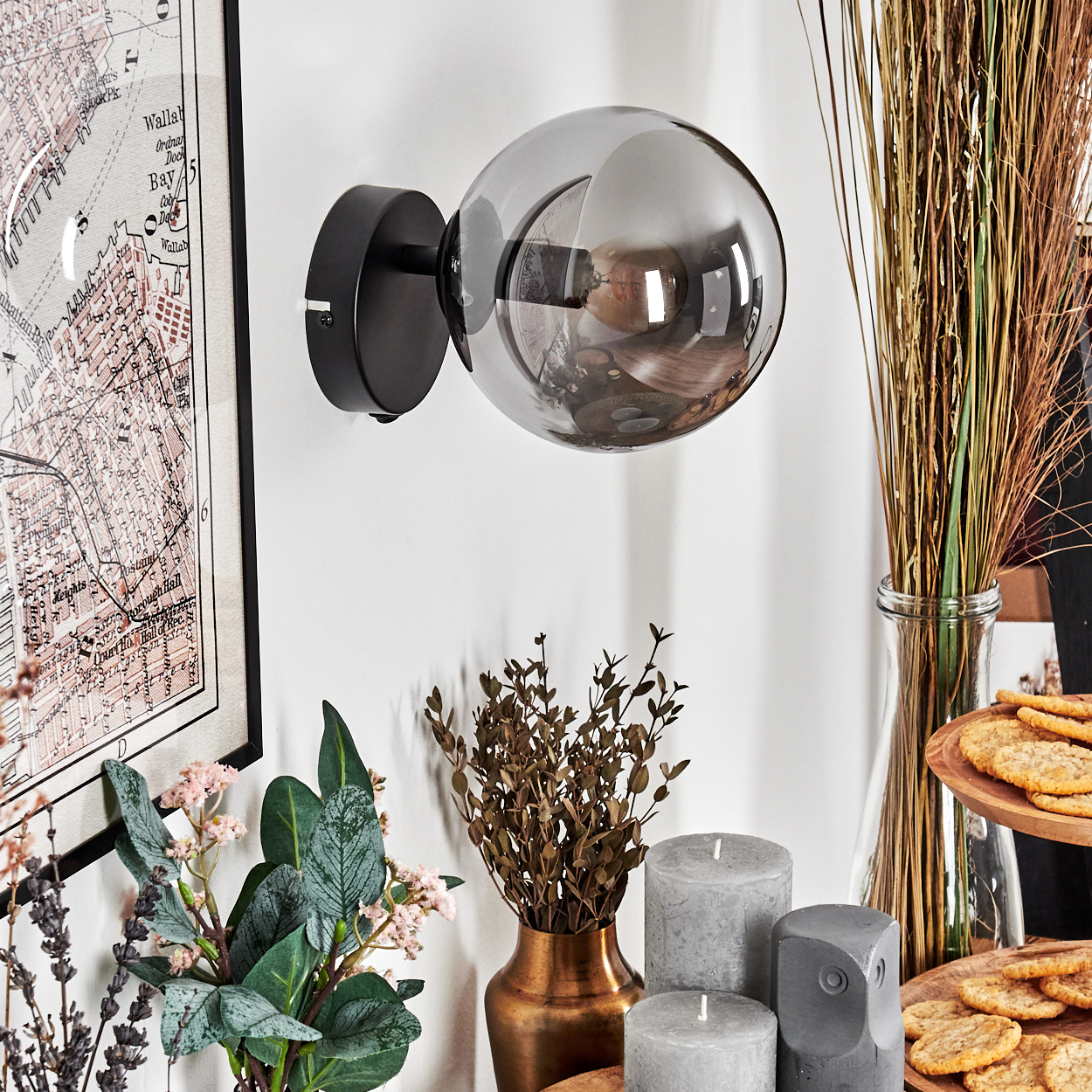 Eine moderne Wandlampe mit einer grauen Glaskugel, Kerzen, getrockneten Pflanzen und einer Karte an der Wand. Ein Tablett mit Keksen ist ebenfalls sichtbar.