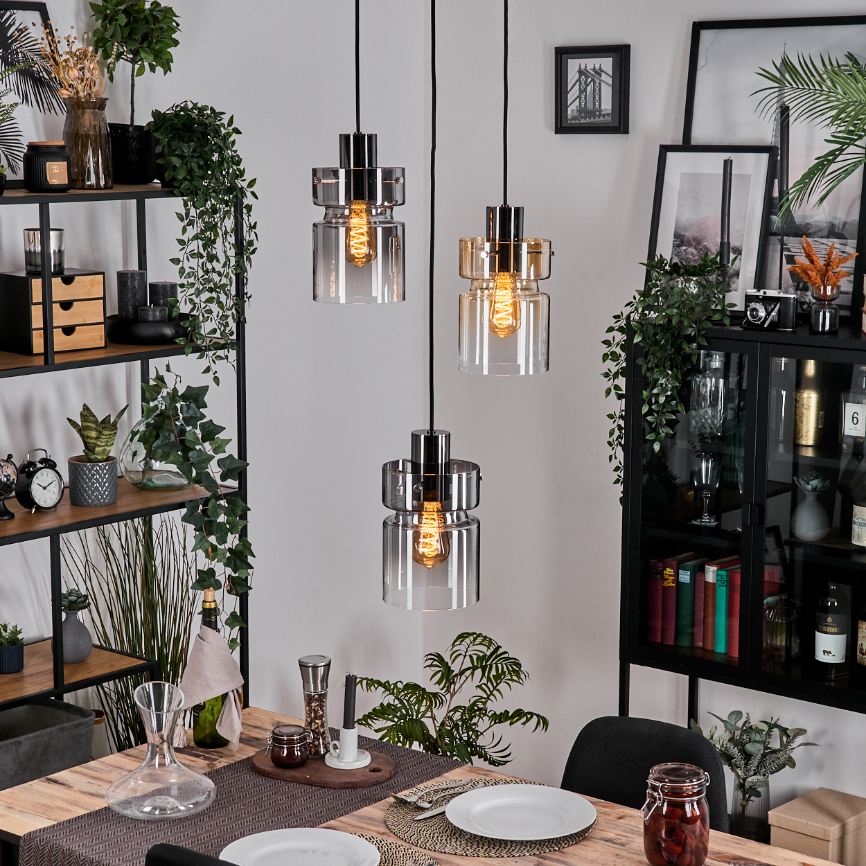 Esszimmer mit Holztisch, drei Hängelampen, Pflanzen und Dekorationsgegenständen auf Regalen und einem Schrank.
