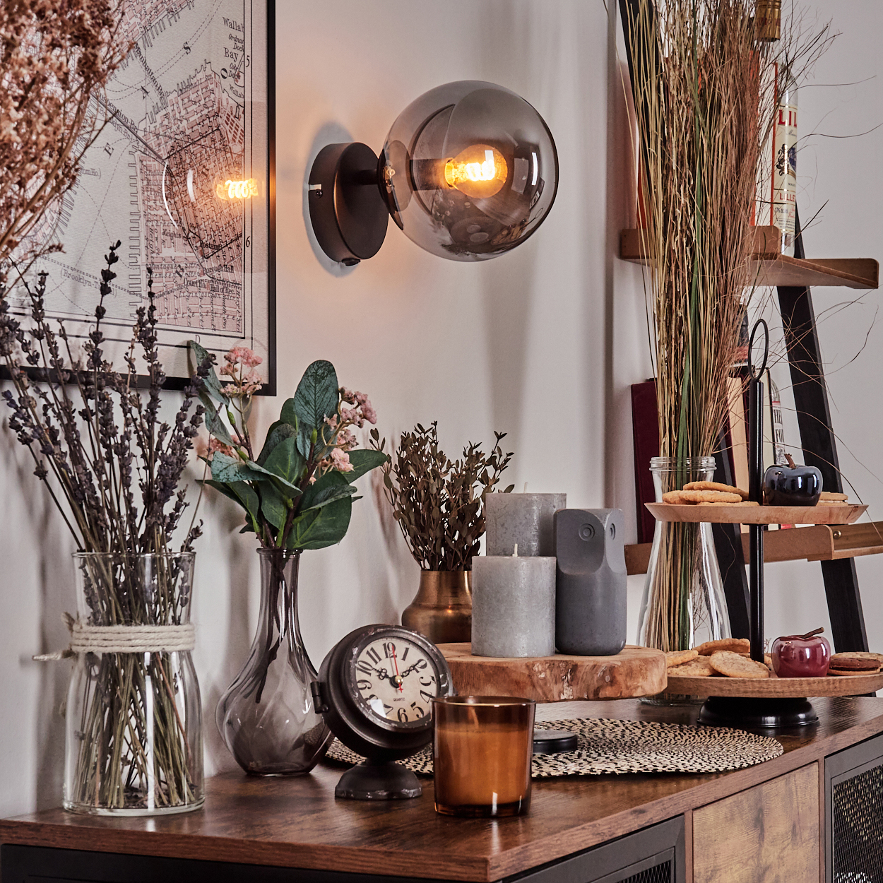 Ein Innenraum mit Wandlampe, Vasen mit Blumen, Uhr, Kerzen und einem Holzregal mit Keksen und Äpfeln.