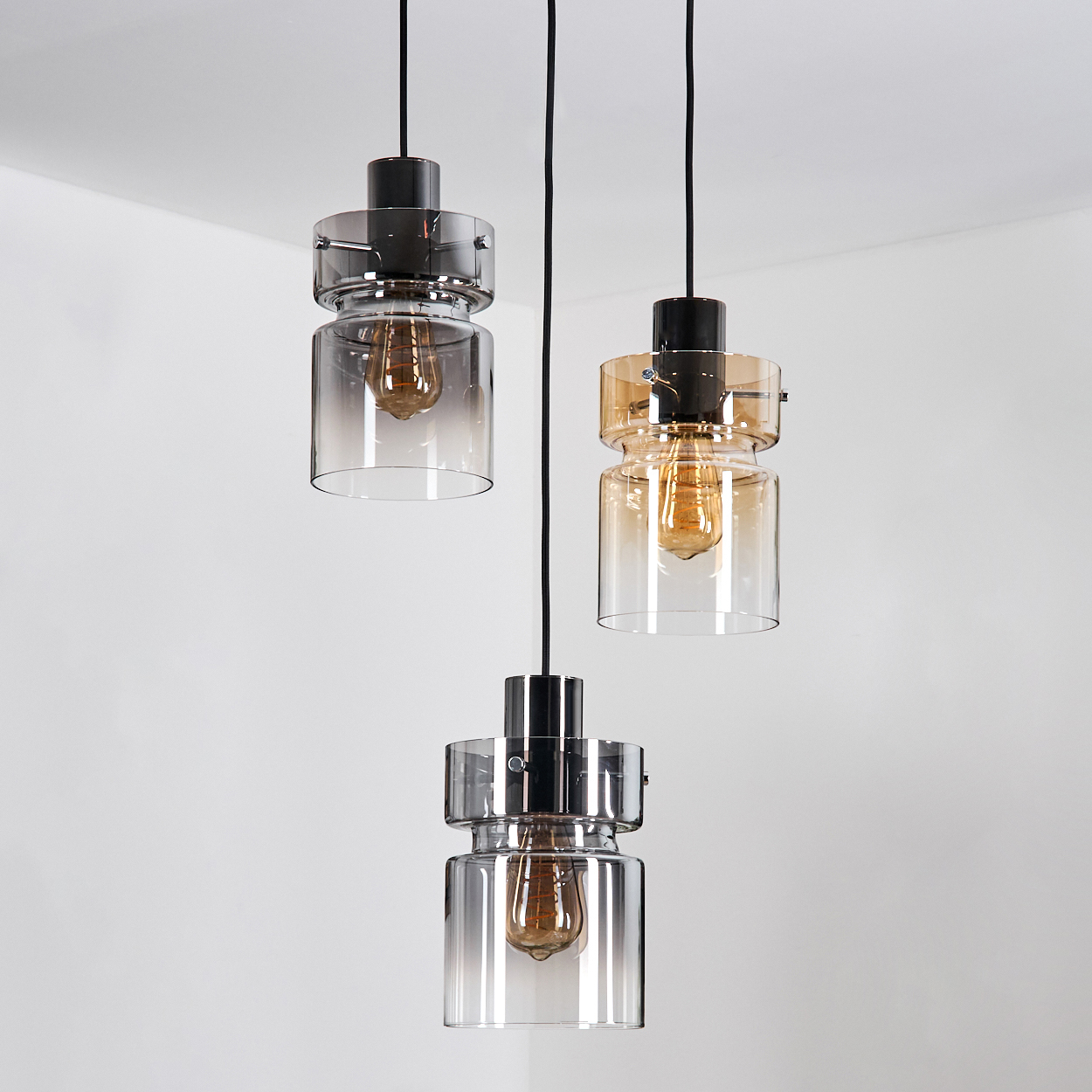 Drei Pendelleuchten mit Glasschirmen und sichtbaren Glühbirnen hängen von der Decke, vor einem schlichten weißen Hintergrund.