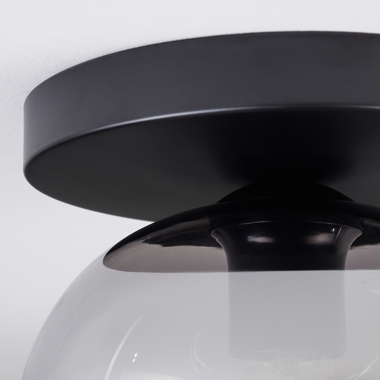 Nahaufnahme einer schwarzen Deckenlampe. Sie hat einen runden Sockel, einen geschwungenen Schirm und eine transparente Glasabdeckung.