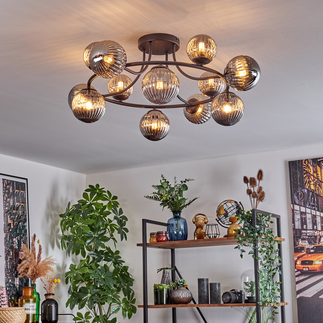 Eine moderne Deckenleuchte mit Glaskugeln hängt über einem Raum mit Pflanzen, Regalen und einem Stadtbildgemälde.