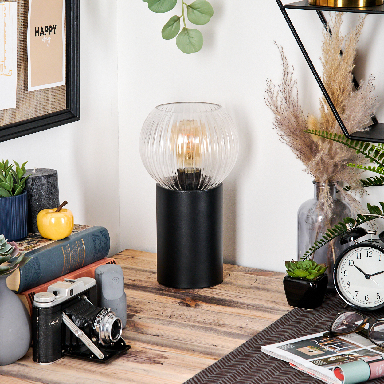 Ein Schreibtisch mit Lampe, Büchern, Kamera, Uhr und anderen Dekorationsartikeln. Der Hintergrund ist eine weiße Wand.