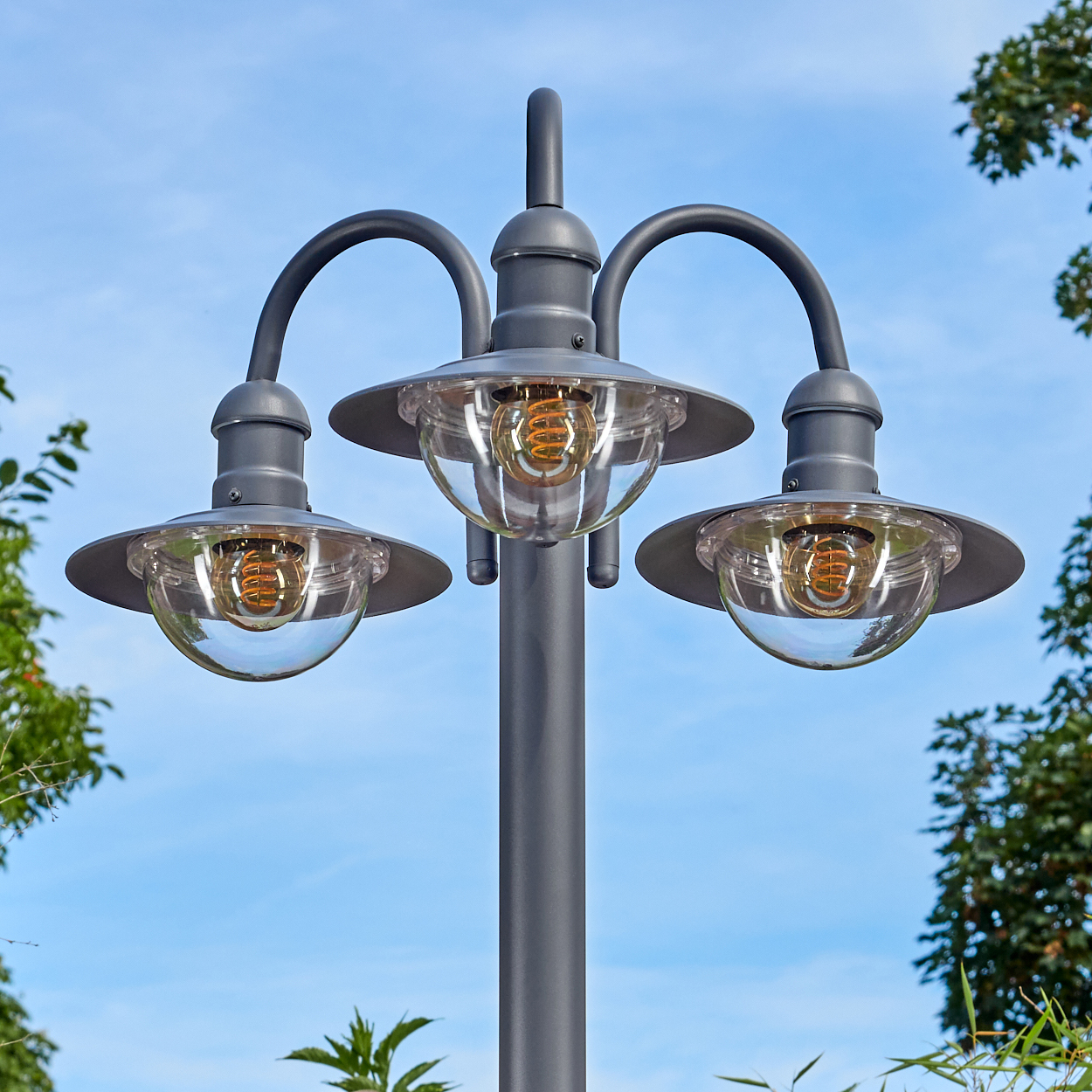 Graue Straßenlaterne mit drei Lichtern, je mit klarem Glasschirm und sichtbarer Glühbirne. Blauer Himmel im Hintergrund.
