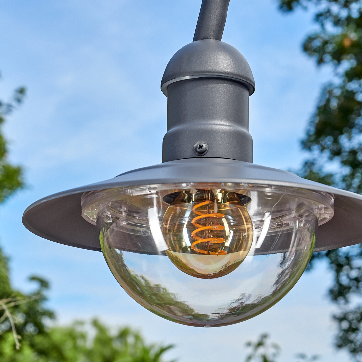 Eine graue Außenlampe mit klarem Glasschirm und warmtoniger Glühbirne, vor blauem Himmel mit grünem Laub.