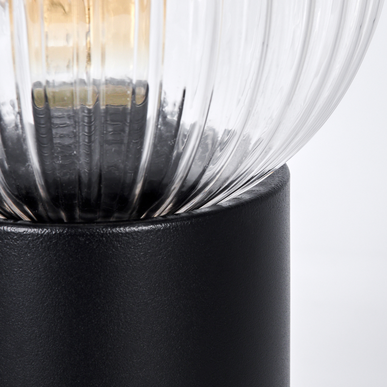 Nahaufnahme einer Lampe. Ein klarer, geriffelter Glasschirm sitzt auf einem schwarzen Sockel. Die Glühbirne ist durch das Glas sichtbar.