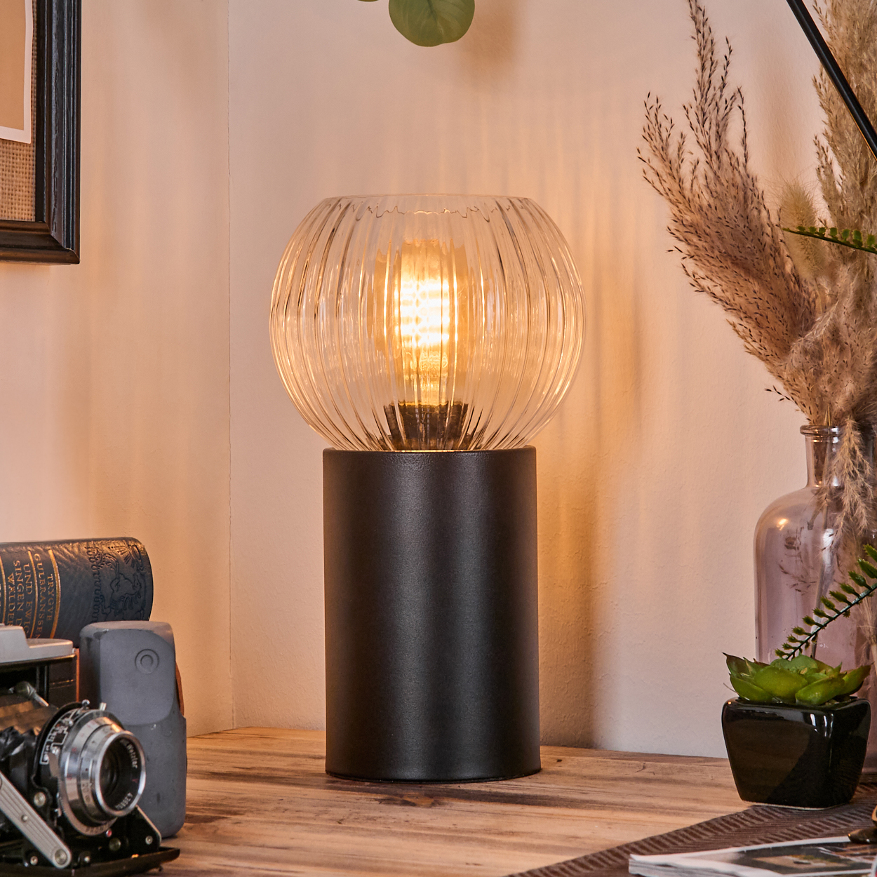 Eine beleuchtete Lampe mit Glasschirm steht auf einem Holztisch. Daneben Bücher, eine Kamera und eine kleine Pflanze. Pampasgras in einer Vase.