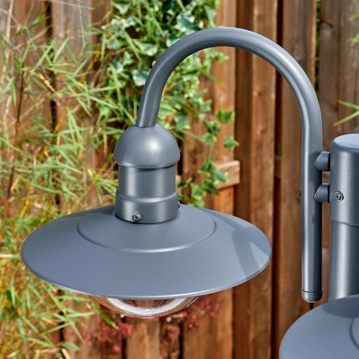 Nahaufnahme einer grauen Außenlampe mit gebogenem Arm. Dahinter ein Holzzaun und grünes Laub.