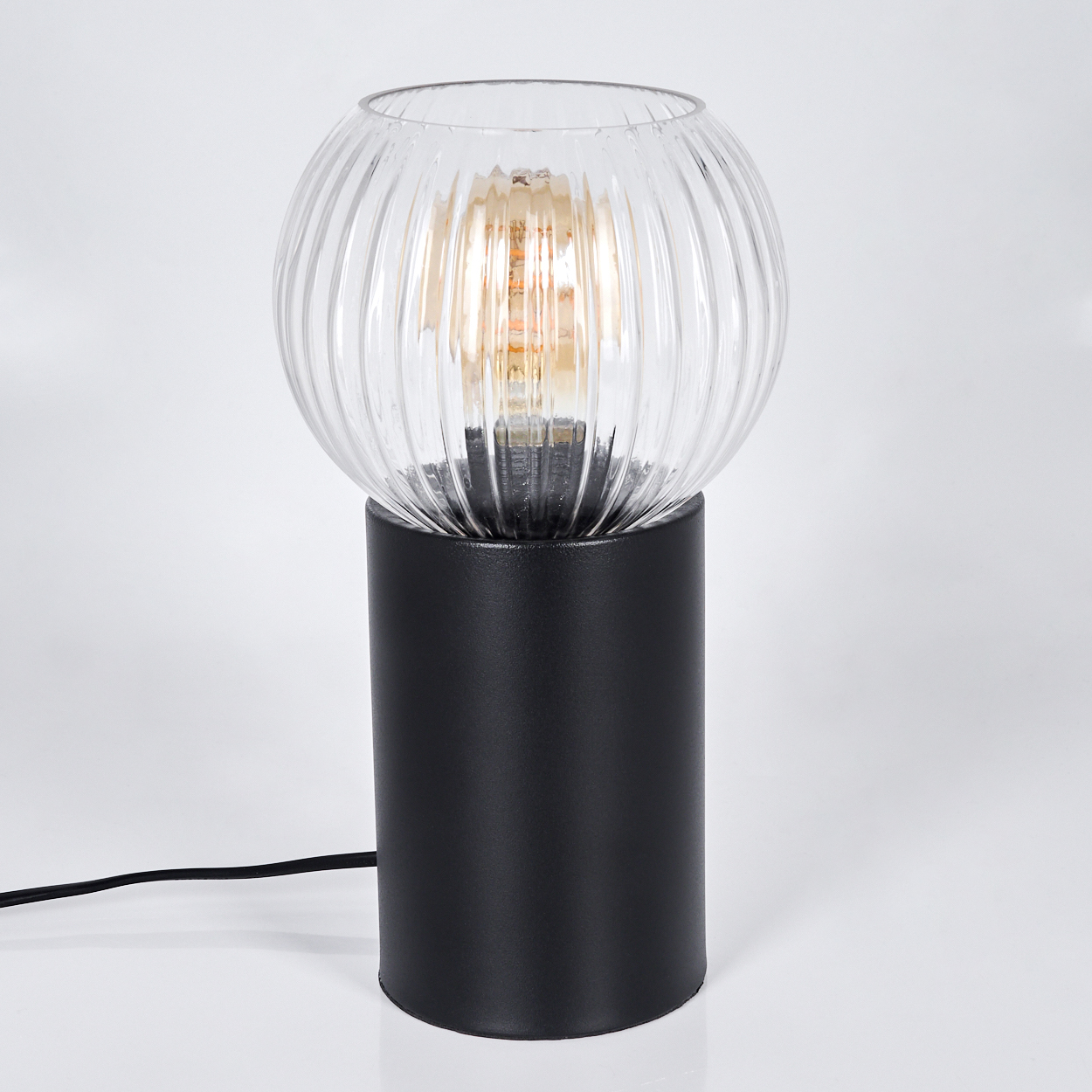 Eine moderne Lampe mit geripptem Glaskugel und zylindrischem schwarzem Sockel. Eine Glühbirne ist im Inneren der Kugel sichtbar. Ein schwarzes Kabel verläuft.