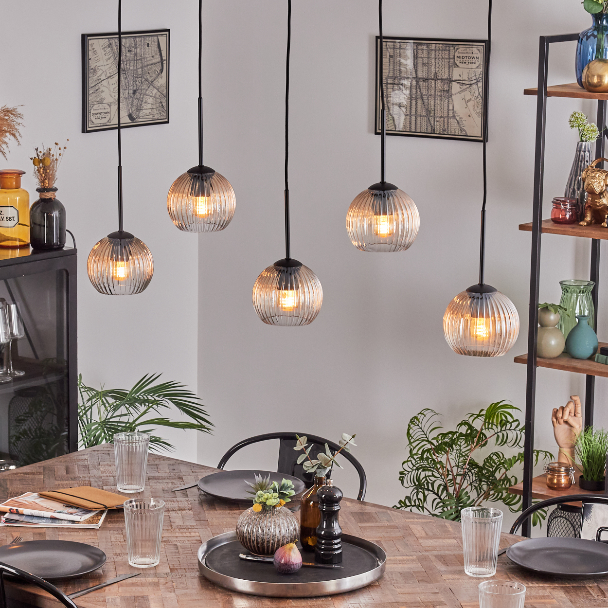 Esszimmer mit Holztisch, fünf Hängelampen, Tellern, Gläsern und Dekoration. Pflanzen und Regale im Hintergrund.