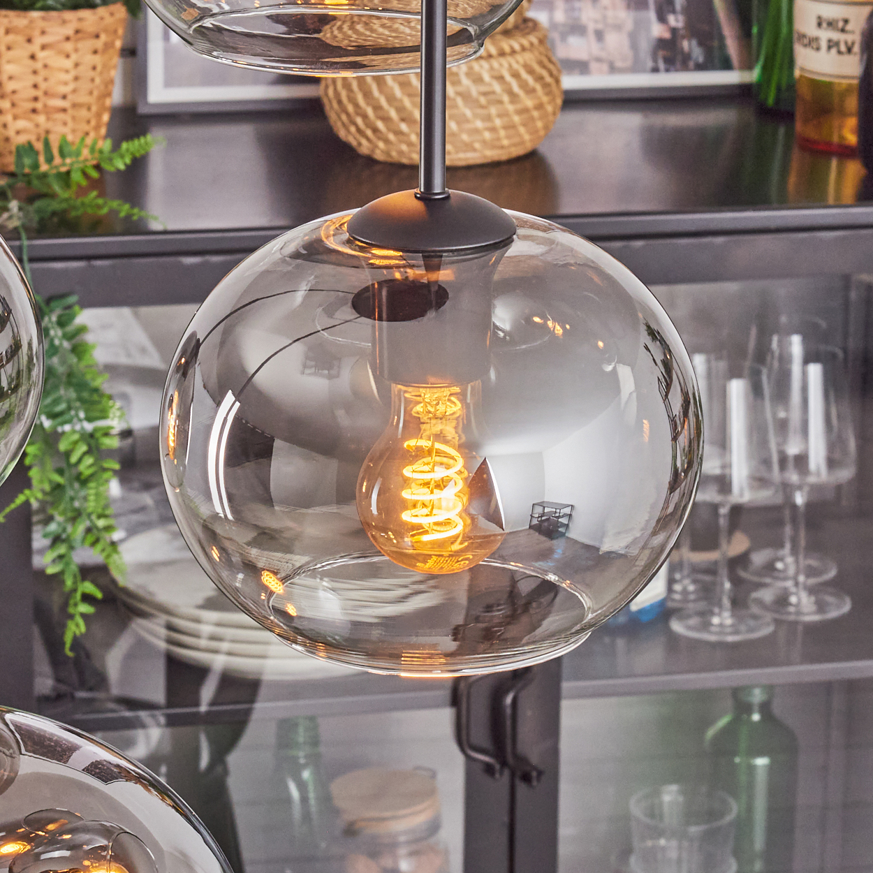 Eine moderne Glas-Pendelleuchte mit leuchtender Wendelglühbirne. Im Hintergrund Regale, Glaswaren und Pflanzen. Warme Beleuchtung.