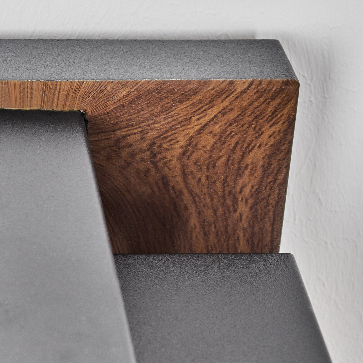 Nahaufnahme einer Holz- und Metallkonstruktion. Das Holz hat eine braune Maserung, das Metall ist grau. Weiße strukturierte Wand im Hintergrund.
