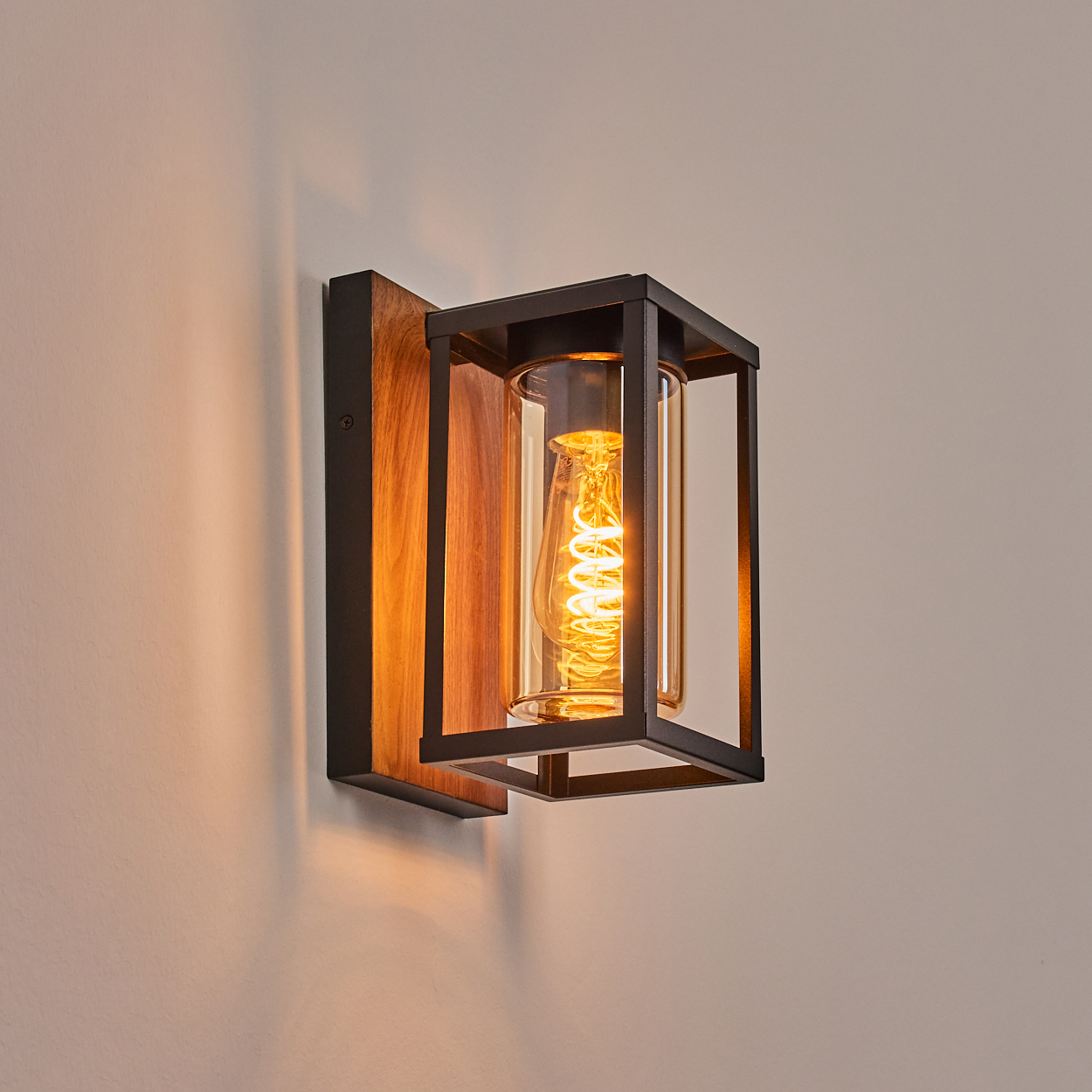 Eine Wandleuchte mit Holzrücken, schwarzem Metallrahmen und einem transparenten Glaszylinder mit einer glühenden Glühbirne.
