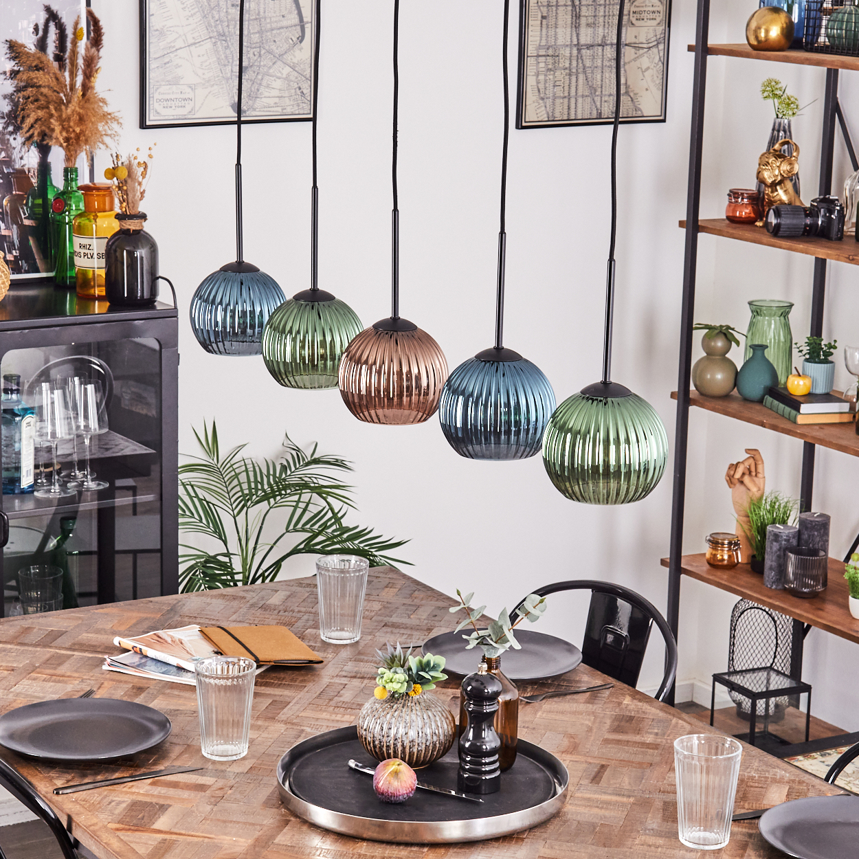 Esszimmer mit Holztisch, Glaslampen, Pflanzen und einem schwarzen Metallregal mit Vasen und Dekoration.
