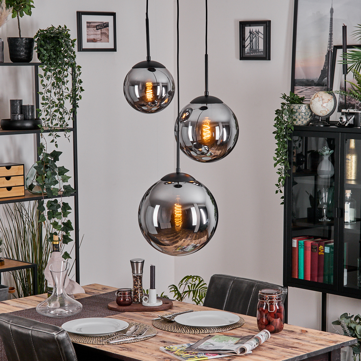 Esszimmer mit Holztisch, drei Hängelampen, Tellern und einem Kirschglas. Regale mit Pflanzen und Dekoration sind sichtbar.