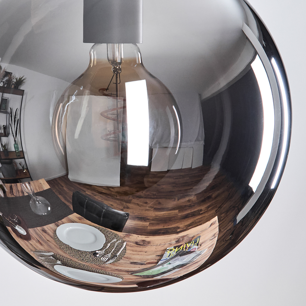 Eine große, reflektierende Glühbirne hängt und spiegelt eine Esszene mit Tisch, Tellern, Besteck und einer Zeitschrift auf einem Holzboden wider.