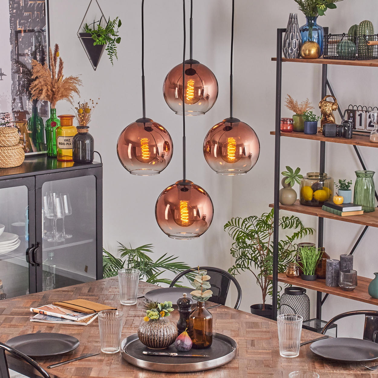 Esszimmer mit Holztisch, vier kupferfarbenen Pendelleuchten und verschiedenen Dekorationsartikeln auf Regalen und dem Tisch.