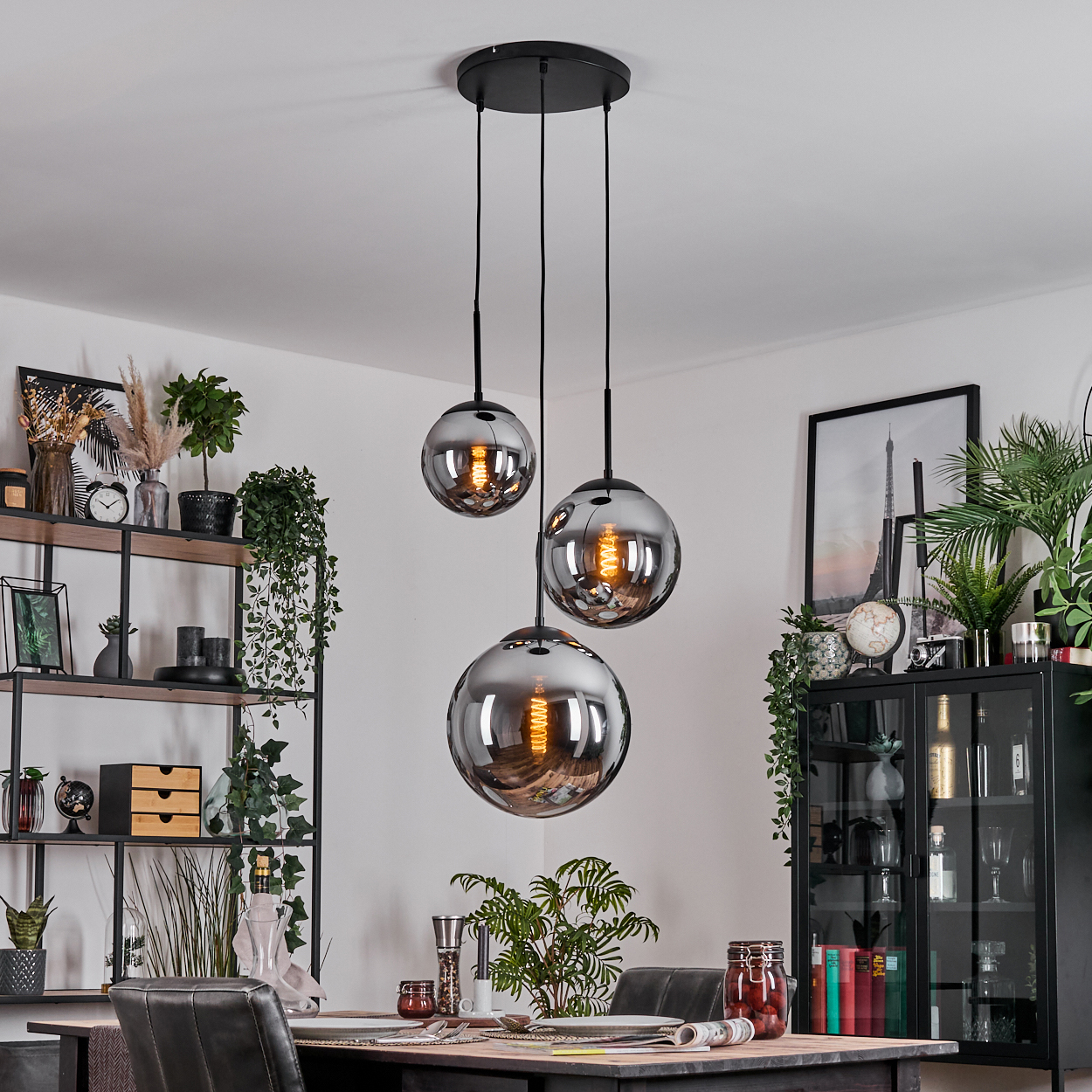 Drei kugelförmige Pendelleuchten hängen über einem Esstisch. Dahinter Regale mit Pflanzen und Dekoration. Ein Schrank und gerahmte Kunst sind sichtbar.