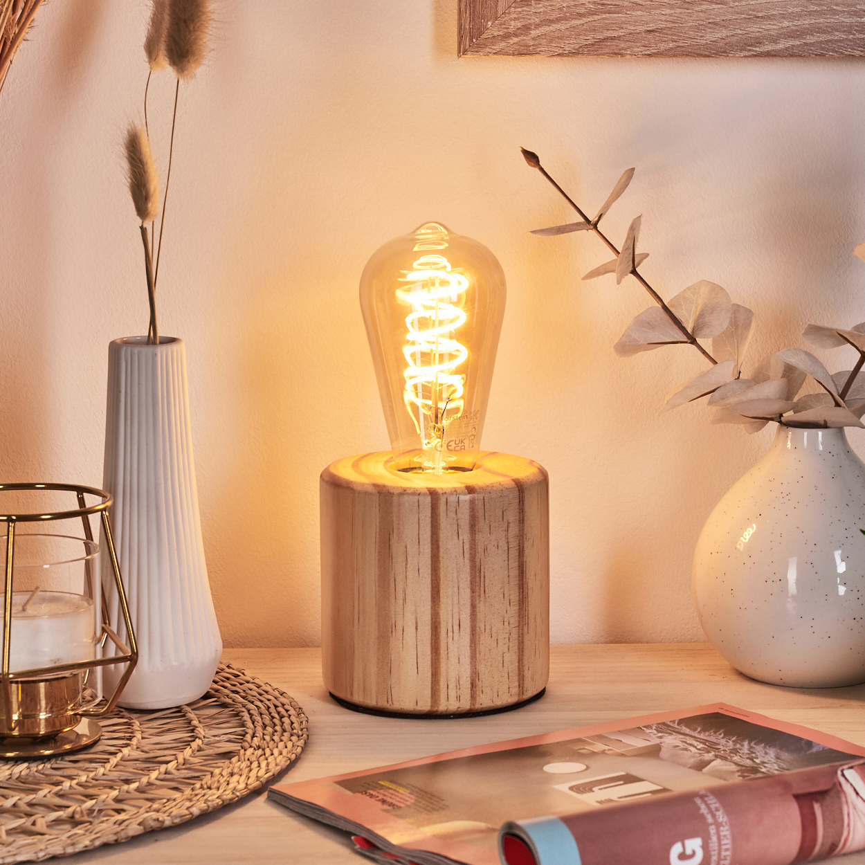 Eine brennende Edison-Gl ühbirnenlampe auf einem Holzsockel. Auf dem Tisch liegen eine Zeitschrift, Vasen mit Pflanzen und eine Kerze.