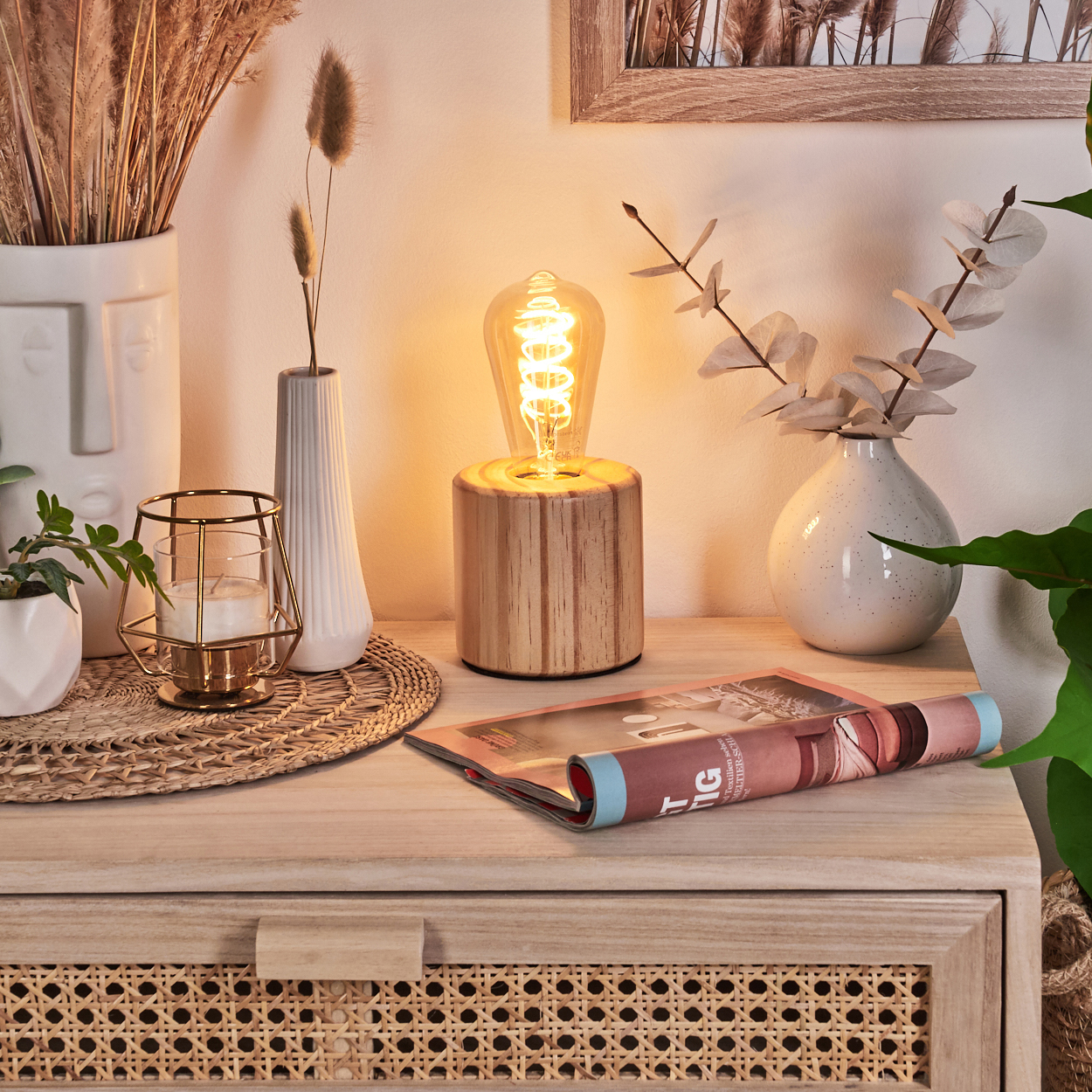 Ein Holztisch mit Lampe, Vase und Zeitschrift. Der Hintergrund hat einen Bilderrahmen.
