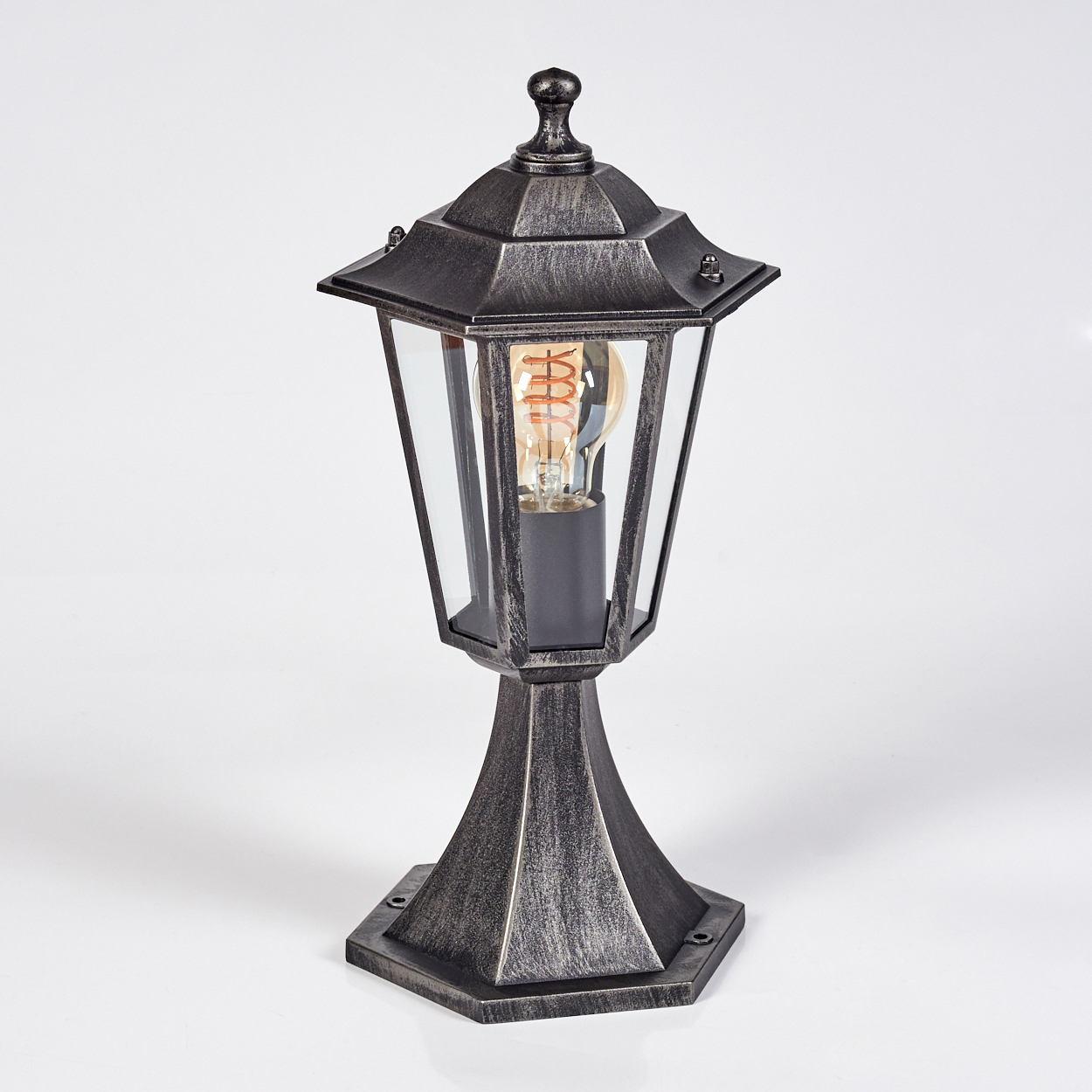 Eine metallgraue Außenlampe mit Glasgehäuse und sichtbarer Glühbirne. Die Lampe hat eine dekorative Oberseite und einen sechseckigen Sockel.