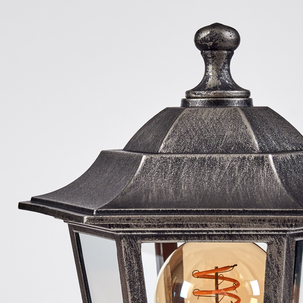 Nahaufnahme einer dekorativen Außenlampe mit silber-grauer Oberfläche und sichtbarer Glühbirne. Der Hintergrund ist weiß.