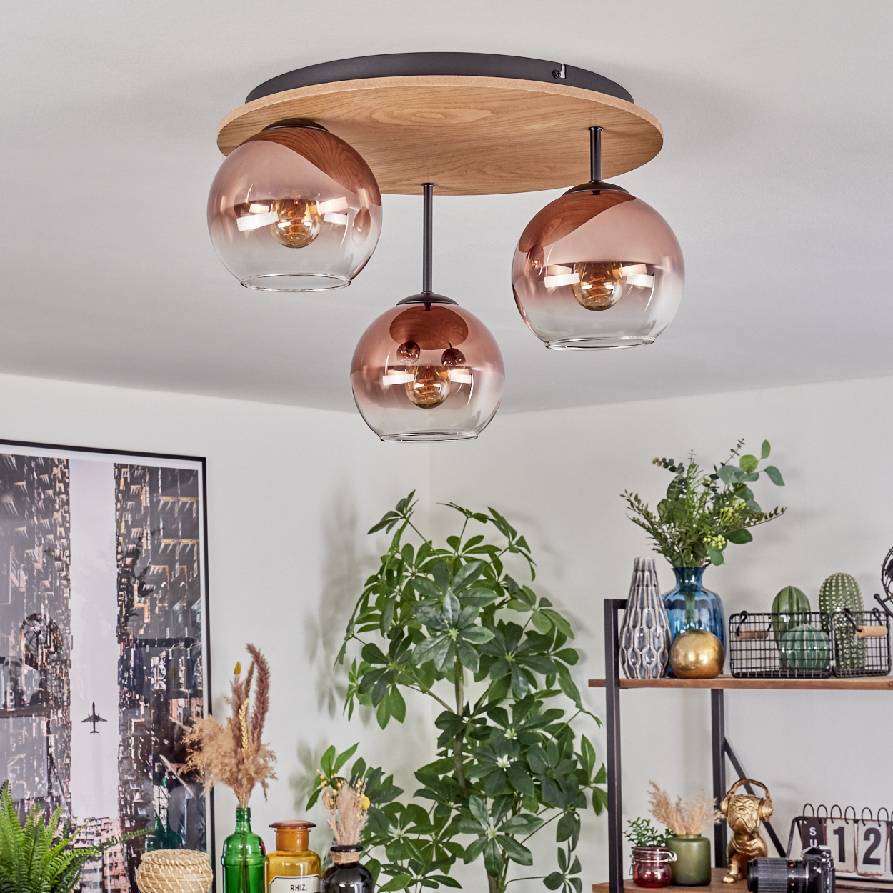 Eine moderne Deckenlampe mit drei kugelförmigen Lampenschirmen hängt über einem Raum mit Pflanzen und Dekorationsgegenständen. Die Schirme sind kupferfarben.