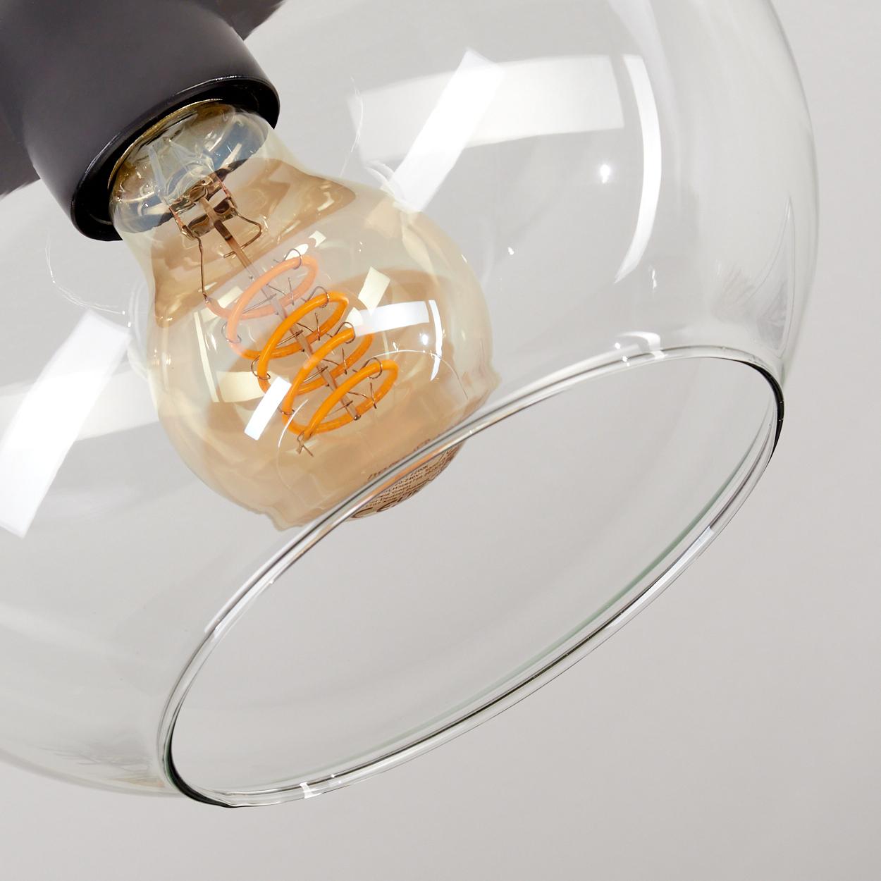 Nahaufnahme einer Glas-Pendelleuchte mit sichtbarer Glühbirne. Die Birne hat einen orangefarbenen Glühfaden. Die Lampe befindet sich auf weißem Hintergrund.