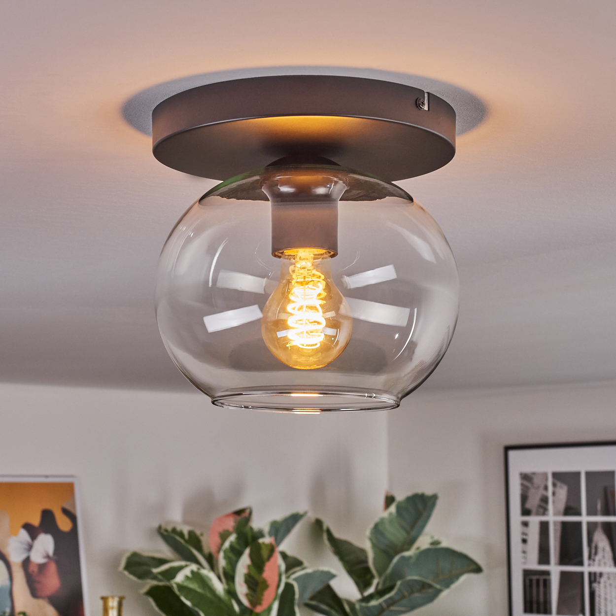 Eine moderne Deckenlampe mit rundem Glasschirm und warmer Glühbirne. Die Leuchte ist auf einem dunklen Metallsockel montiert.